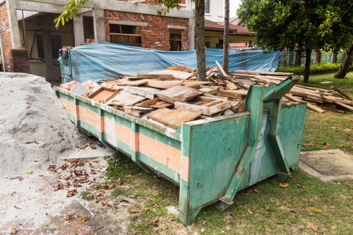 A large, green metal skip positioned on a patch of grass near a residential area, filled with a variety of discarded wooden planks, panels, and boards of different sizes, some with visible nails and rough edges. The wooden materials display a mixture of natural wood tones, from light beige to darker brown, with some showing weathered or worn surfaces. Behind the skip, a suburban street scene is visible with a partially constructed or renovation-in-progress brick house, protected by a blue tarpaulin sheet stretched across scaffolding. The scene is shaded by nearby trees with green foliage, and there are fallen leaves scattered around the base of the skip, indicating an outdoor environment. This image, associated with Commercial Waste Removal Carshalton, illustrates typical rubbish clearance involving wooden waste in a domestic or small commercial context, suitable for services in the local postcode area.