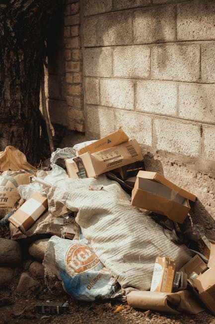 A street scene showing a large, white refuse collection truck with visible rust and mechanical components on the rear. The truck is positioned parallel to an older, multi-storey building with resident
