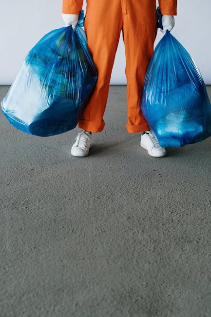 A person standing indoors on a grey carpeted floor, wearing orange work overalls and white sneakers, holding a large blue plastic rubbish bag in each hand. The bags appear filled with waste, with visi